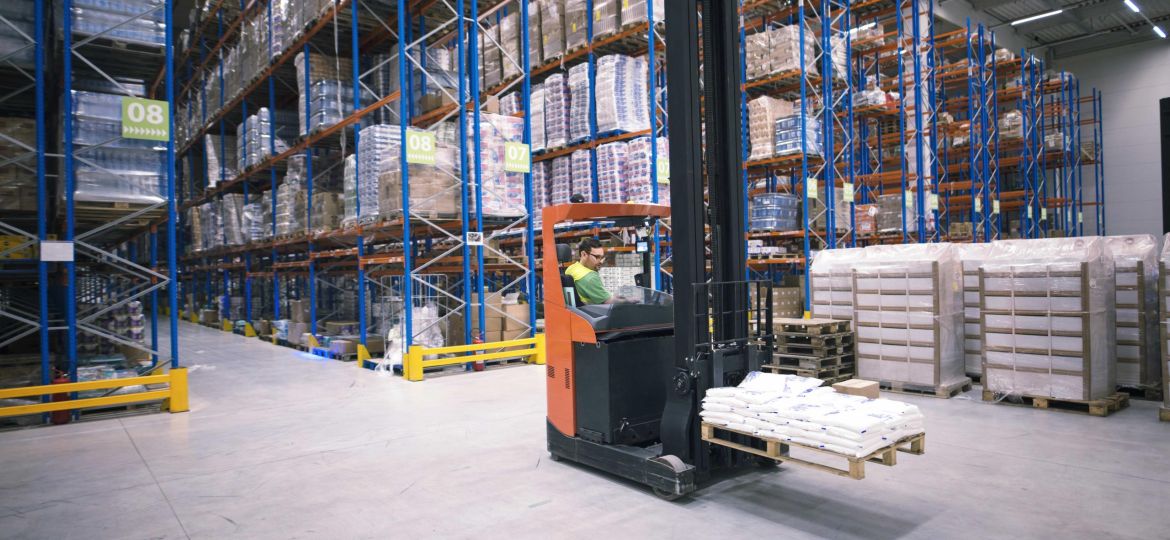 Worker operating forklift machine and relocating goods in large warehouse center.